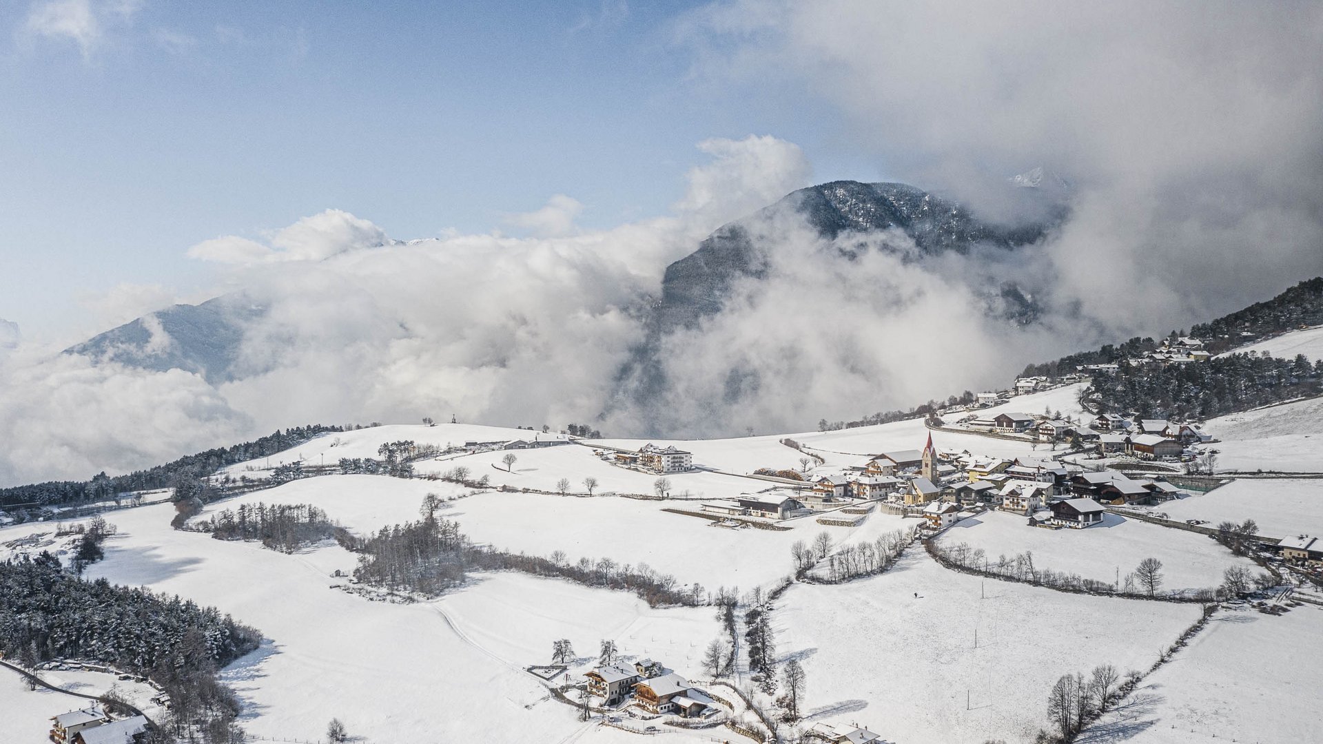 La frazione montana di Spinga L'immagine mostra un villaggio innevato situato in un paesaggio dolcemente collinare. Le montagne sullo sfondo sono parzialmente coperte da nuvole, e il cielo è di un azzurro limpido, rendendo il paesaggio invernale luminoso e pacifico.
