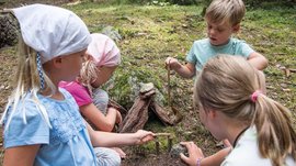 Il Sentiero degli Elfi a Vandoies di Sopra L'immagine mostra quattro bambini che giocano insieme nella foresta. Stanno costruendo una piccola struttura con bastoni, pietre e corteccia sul terreno, circondati da muschio e alberi.