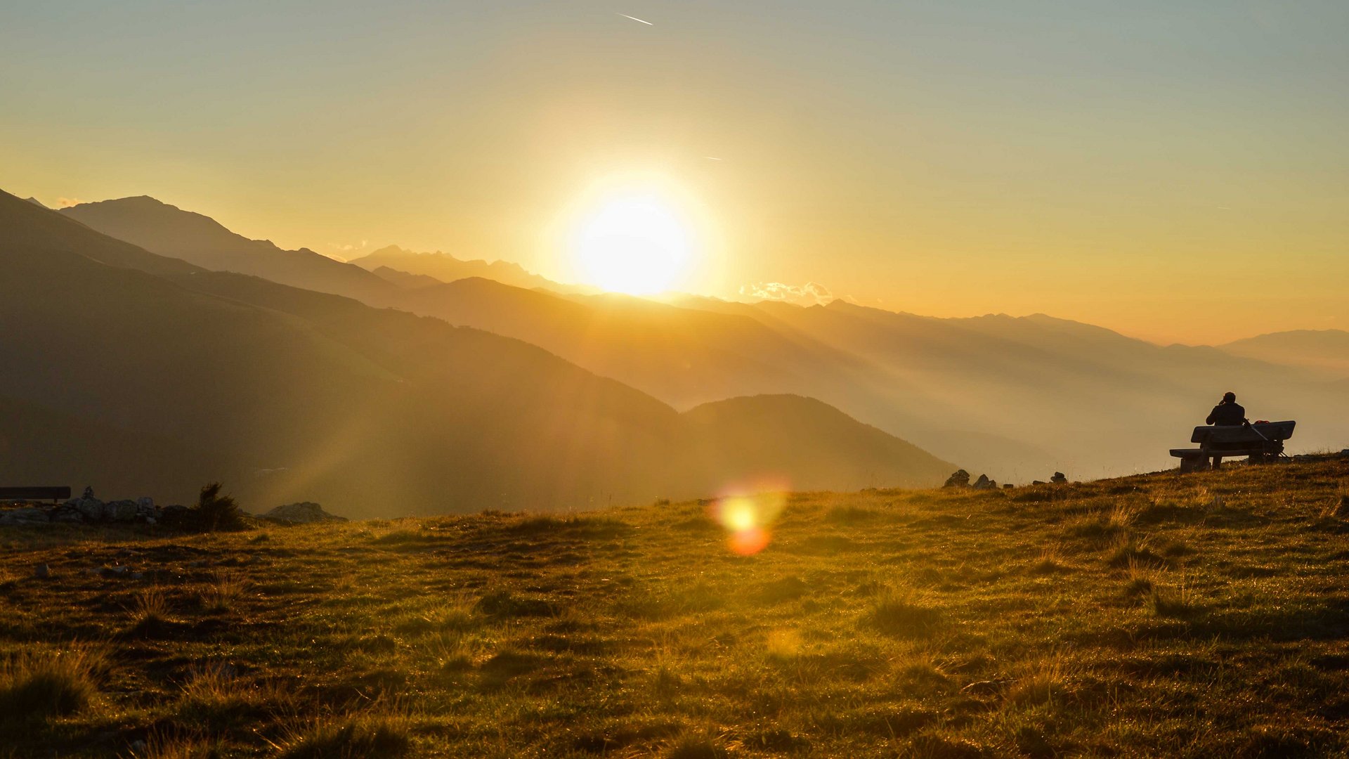 Valles - Jochtal - Steinermandl L'immagine mostra un tramonto su un paesaggio montano, con il sole che si trova basso sopra le vette delle montagne e proietta una luce dorata e delicata sul prato. Su una panchina in primo piano siede una persona che guarda verso il sole al tramonto.