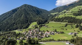 Quando si dice idilliaco… ecco a voi Fundres L'immagine mostra un idilliaco paesaggio verde con un piccolo villaggio che si estende in una valle tra pendii montuosi boscosi. Le case del villaggio sono circondate da prati rigogliosi e, sullo sfondo, si erge una montagna densamente boscosa sotto un cielo azzurro con qualche nuvola bianca.