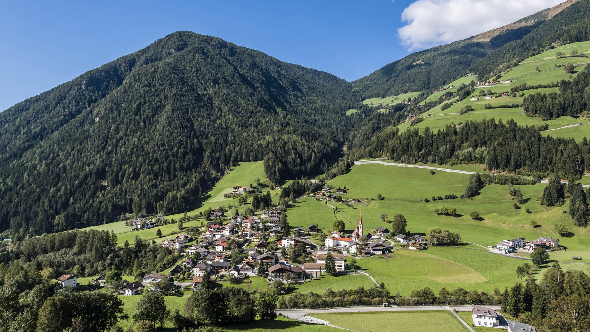 Quando si dice idilliaco… ecco a voi Fundres L'immagine mostra un idilliaco paesaggio verde con un piccolo villaggio che si estende in una valle tra pendii montuosi boscosi. Le case del villaggio sono circondate da prati rigogliosi e, sullo sfondo, si erge una montagna densamente boscosa sotto un cielo azzurro con qualche nuvola bianca.