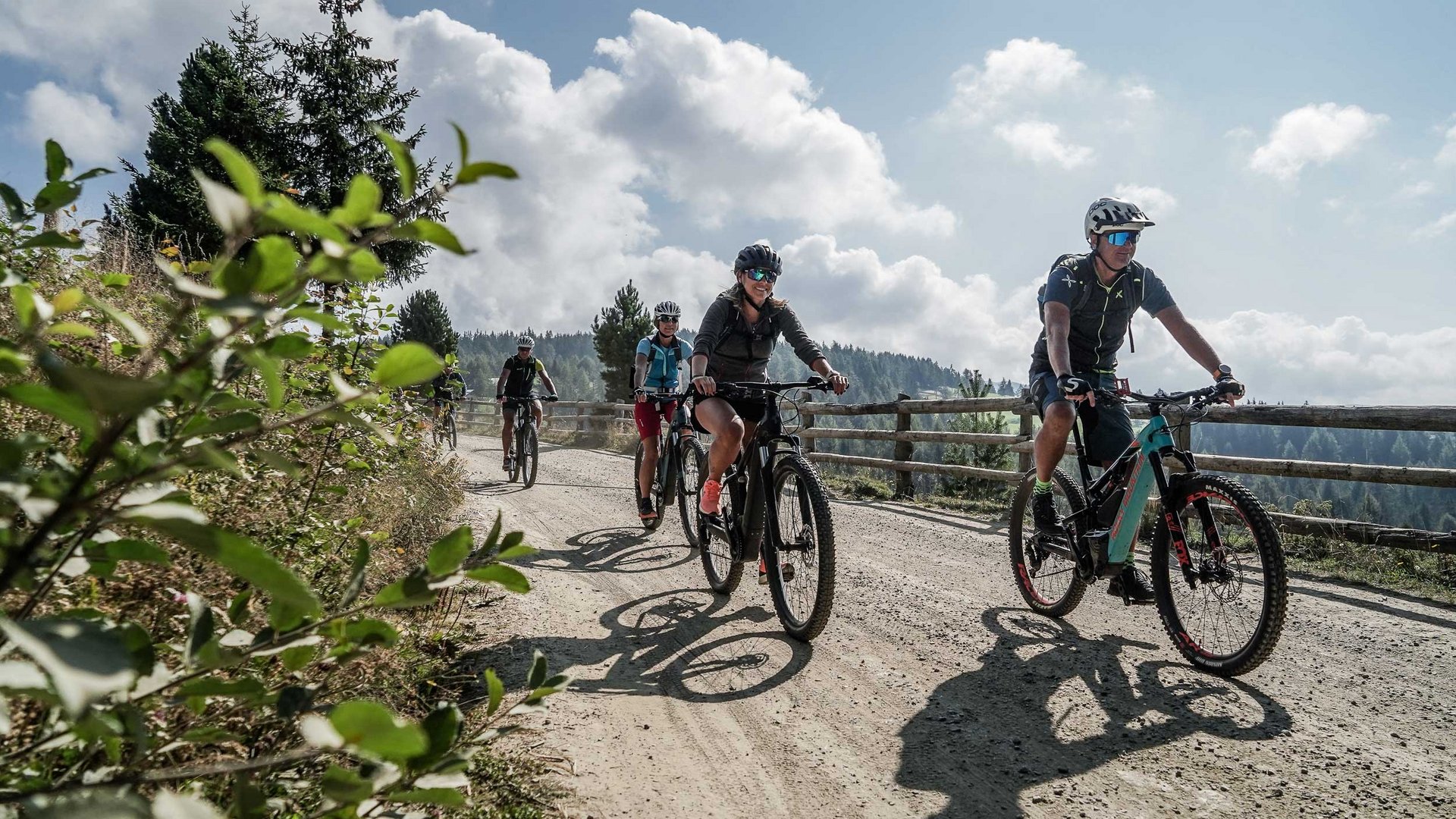 Le Settimane Bike in Alto Adige L'immagine mostra un gruppo di ciclisti che percorre un sentiero forestale soleggiato. I ciclisti indossano caschi e abbigliamento sportivo, mentre sullo sfondo si vedono alberi e un cielo nuvoloso.