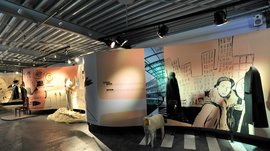 Il Mondo del Loden a Vandoies L'immagine mostra un'installazione museale in cui sono esposti abiti storici. Sulle pareti si vedono illustrazioni di scene di diverse epoche, tra cui una vista della città. Davanti alle pareti ci sono manichini vestiti con cappotti e altri indumenti, oltre a un modello di una pecora sul pavimento.