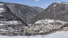 Alla scoperta di Rio di Pusteria L'immagine mostra una città situata in una valle innevata, circondata da colline densamente boscose. Gli edifici sono coperti di neve e sullo sfondo si vedono colline innevate e un cielo azzurro e limpido.
