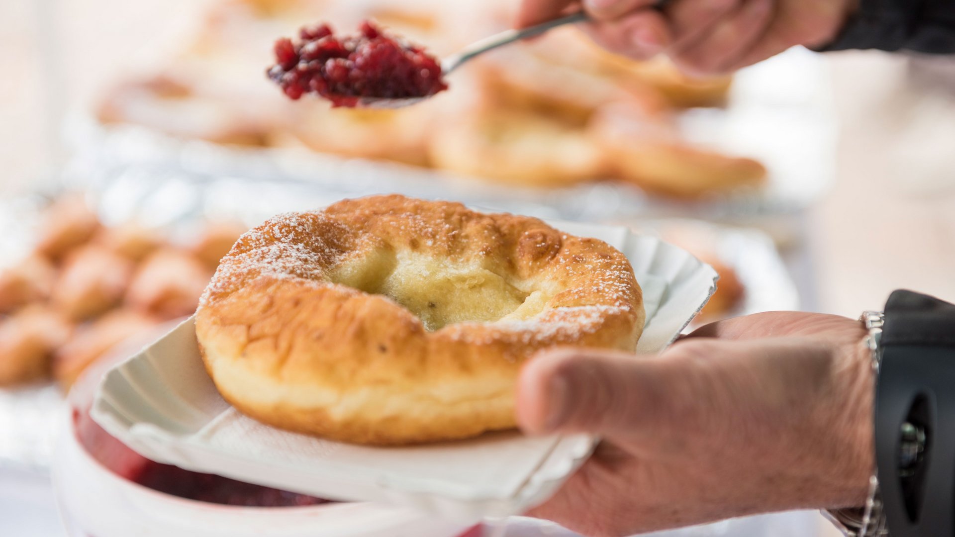 Shopping a Rio di Pusteria (BZ) L'immagine mostra una persona che sta mettendo con un cucchiaio una marmellata rossa o una composta su un dolce fritto, tenuto in mano su un piatto di carta. Il dolce è spolverato con zucchero a velo e appare dorato e croccante.