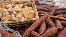 Shopping a Rio di Pusteria (BZ) L'immagine mostra un cesto con pezzi di pane tagliati in modo rustico. Accanto al cesto ci sono numerosi insaccati speziati e stagionati all'aria in un'altra cassetta intrecciata. La scena suggerisce un ambiente tradizionale di mercato o fattoria.