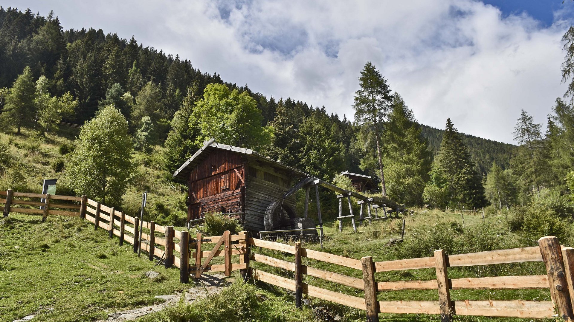 Il Sentiero dei Mulini a Terento Casa di legno con ruota ad acqua su prato verde con foresta e cielo blu