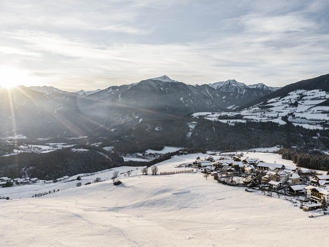 Le località dell’area vacanze Rio Pusteria L'immagine mostra un paesaggio innevato nelle montagne, poco dopo l'alba. Il sole sorge dietro le montagne e illumina la valle, caratterizzata da campi e colline ricoperte di neve. Sul lato destro dell'immagine si trova un piccolo villaggio, anch'esso coperto di neve. Le montagne sullo sfondo sono in parte boscose e visibili in lontananza.