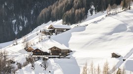 Quando si dice idilliaco… ecco a voi Fundres L'immagine mostra un paesaggio montano invernale con colline innevate e diverse baite distribuite sui pendii. Sullo sfondo si vede una foresta fitta, e la luce del sole basso all'orizzonte proietta lunghe ombre sulla neve.