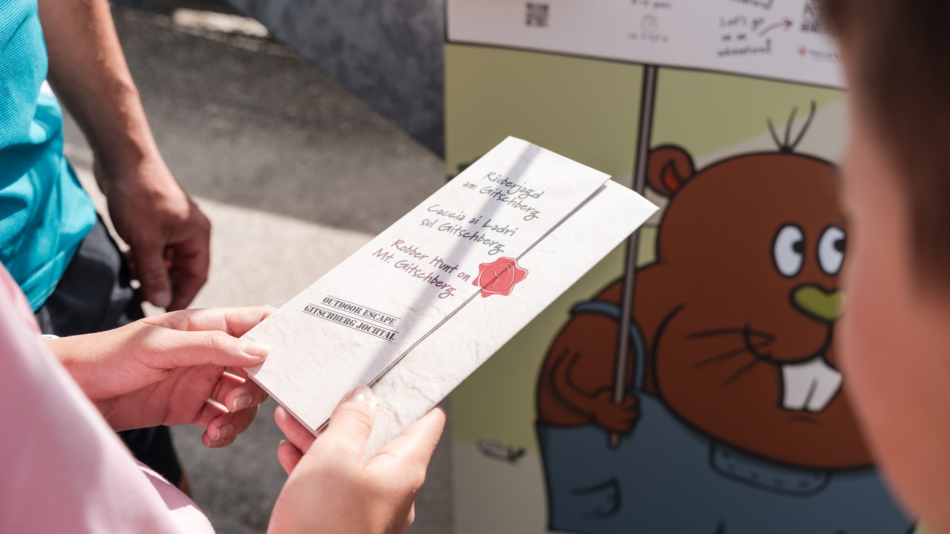 Outdoor Escape sul Monte Gitschberg Persona tiene volantino per caccia ai ladri sul Gitschenberg davanti a tabellone colorato