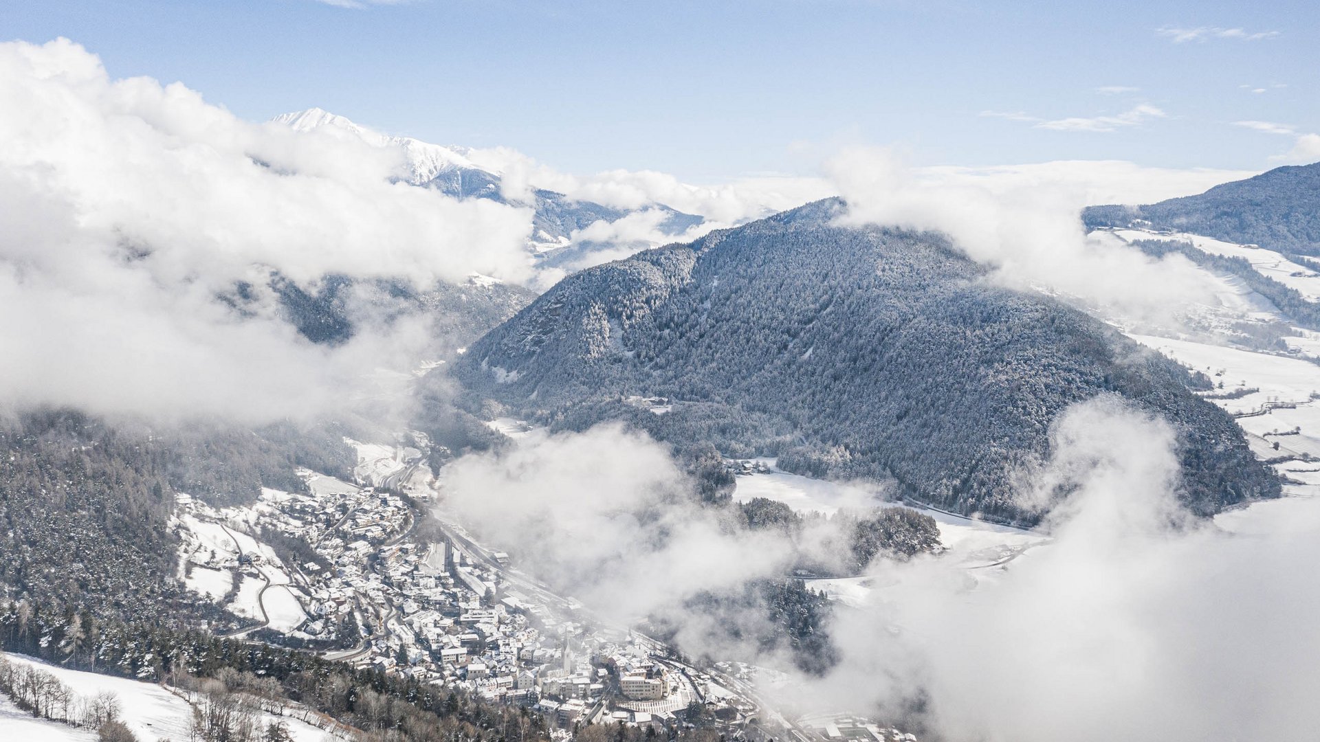 Alla scoperta di Rio di Pusteria L'immagine mostra un paesaggio innevato con una valle e una piccola città circondata da colline boschive e montagne. Nuvole scorrono sulla scena, coprendo parte del paesaggio e creando un'impressione pittoresca e invernale.