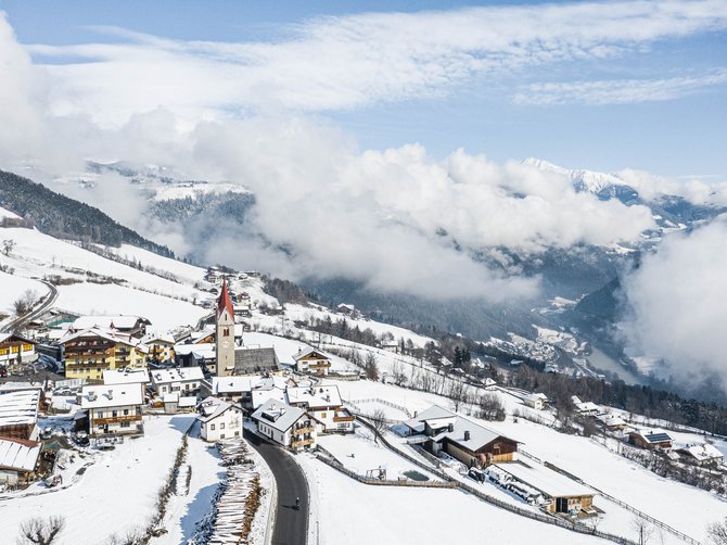 Le località dell’area vacanze Rio Pusteria L'immagine mostra un villaggio di montagna innevato con una chiesa, la cui torre rossa spicca tra le case coperte di neve. Sullo sfondo si vedono colline boschive e nuvole basse che coprono parzialmente la valle sottostante.