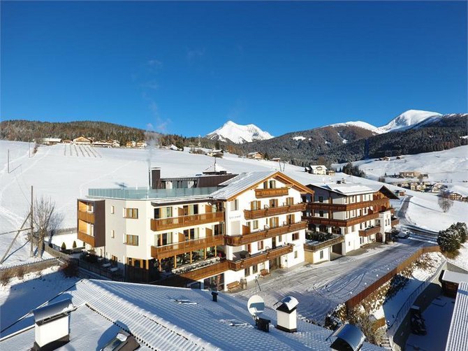 Benvenuti nell’Area Vacanze Sci e Malghe Rio Pusteria! L'immagine mostra un grande hotel in stile alpino, circondato da colline e montagne innevate. Il paesaggio sullo sfondo è composto da campi e boschi innevati sotto un cielo limpido e di un azzurro brillante.