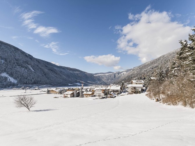 Le località dell’area vacanze Rio Pusteria L'immagine mostra un paesaggio innevato con un piccolo villaggio nella valle, circondato da montagne coperte di neve. Il cielo è limpido e blu, attraversato solo da qualche nuvola, e in primo piano c'è un albero spoglio sul campo innevato.