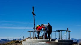 La piattaforma panoramica Gitschberga a Maranza (BZ) L'immagine mostra due escursionisti sulla cima di una montagna che guardano attraverso un binocolo fisso. Sullo sfondo si vede una grande croce di vetta e il cielo è limpido e azzurro.