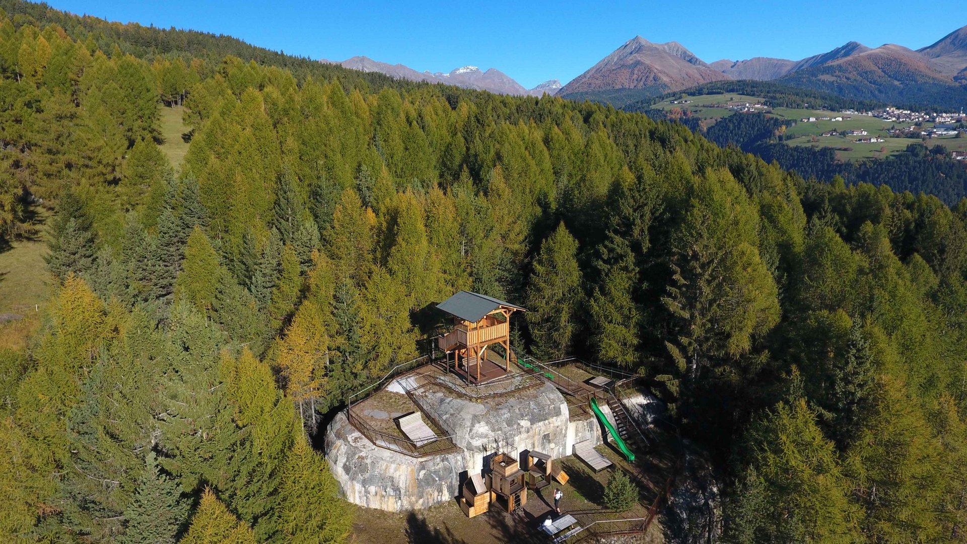 La zona ricreativa bunker di Spinga L'immagine mostra una piattaforma panoramica in legno situata su un promontorio roccioso nel mezzo di una fitta foresta. Sullo sfondo si vedono montagne verdi e un cielo azzurro limpido, che offrono una vista ampia sul paesaggio.