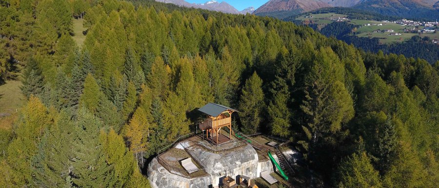 Le località dell’area vacanze Rio Pusteria L'immagine mostra una piattaforma panoramica in legno situata su un promontorio roccioso nel mezzo di una fitta foresta. Sullo sfondo si vedono montagne verdi e un cielo azzurro limpido, che offrono una vista ampia sul paesaggio.