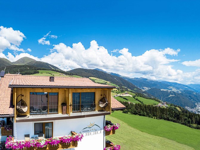 Benvenuti nell’Area Vacanze Sci e Malghe Rio Pusteria! L'immagine mostra un grande edificio con balconi decorati con fiori, davanti a un pittoresco paesaggio montano. Sullo sfondo si vedono colline verdi, foreste e la valle che si estende sotto un cielo azzurro brillante.