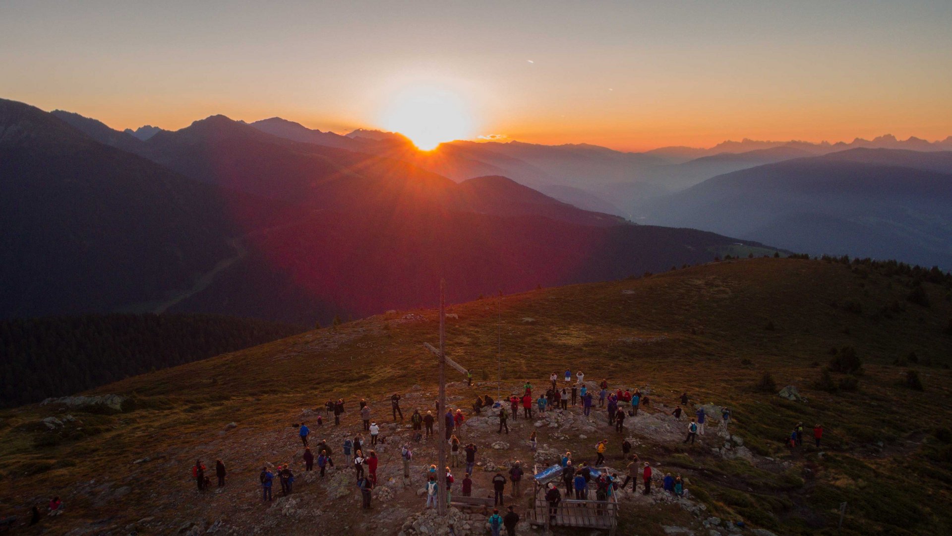 Valles - Jochtal - Steinermandl L'immagine mostra un tramonto su un paesaggio montano, con numerose persone radunate su una vetta. Al centro dell'immagine si trova una grande croce di vetta, mentre il cielo e le montagne brillano in tonalità calde di arancione e rosso.