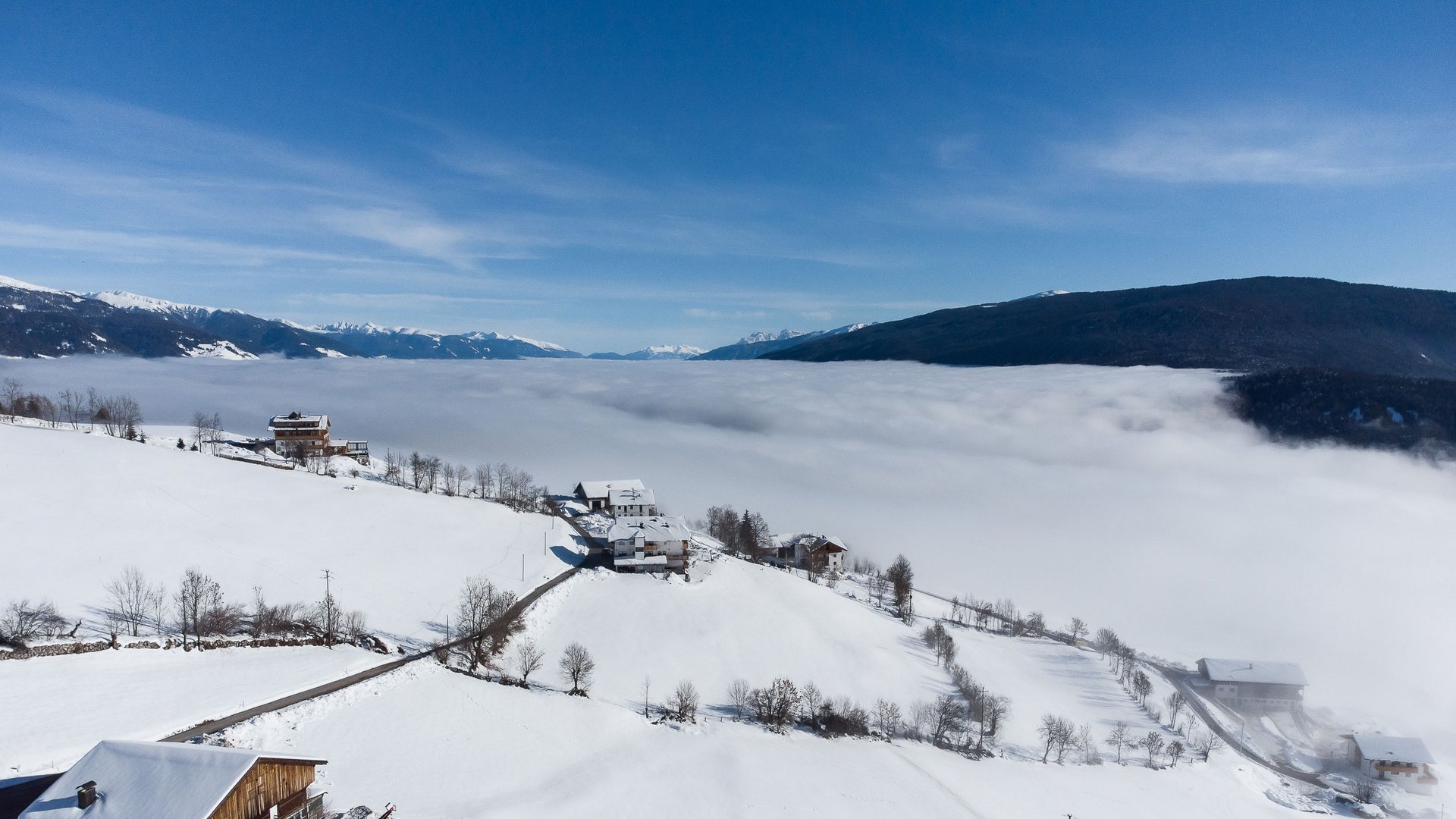 Conoscete già Maranza? L'immagine mostra un paesaggio montano invernale con campi coperti di neve e case sparse su una collina. Sullo sfondo si estende un denso mare di nuvole sopra la valle, mentre le cime delle montagne innevate emergono sotto un cielo limpido e azzurro.