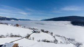 Conoscete già Maranza? L'immagine mostra un paesaggio montano invernale con campi coperti di neve e case sparse su una collina. Sullo sfondo si estende un denso mare di nuvole sopra la valle, mentre le cime delle montagne innevate emergono sotto un cielo limpido e azzurro.