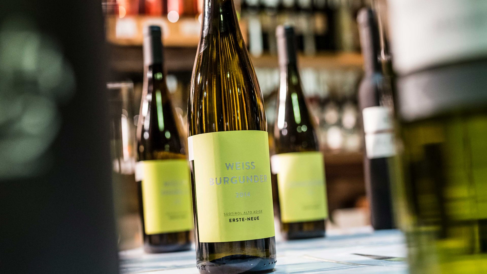 Shopping a Rio di Pusteria (BZ) L'immagine mostra una bottiglia di vino Pinot bianco con un'etichetta gialla, su cui è scritto "Weiss Burgunder 2014". Sullo sfondo si intravedono altre bottiglie di vino sfocate, anch'esse posizionate su uno scaffale.