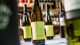 Shopping a Rio di Pusteria (BZ) L'immagine mostra una bottiglia di vino Pinot bianco con un'etichetta gialla, su cui è scritto "Weiss Burgunder 2014". Sullo sfondo si intravedono altre bottiglie di vino sfocate, anch'esse posizionate su uno scaffale.