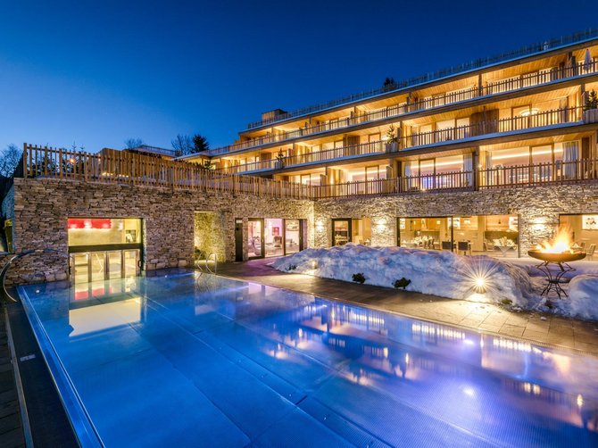 Benvenuti nell’Area Vacanze Sci e Malghe Rio Pusteria! L'immagine mostra un hotel illuminato al crepuscolo con grandi finestre e terrazze su più piani. In primo piano c'è una piscina illuminata, circondata da superfici coperte di neve e da una ciotola di fuoco che crea un'atmosfera accogliente.