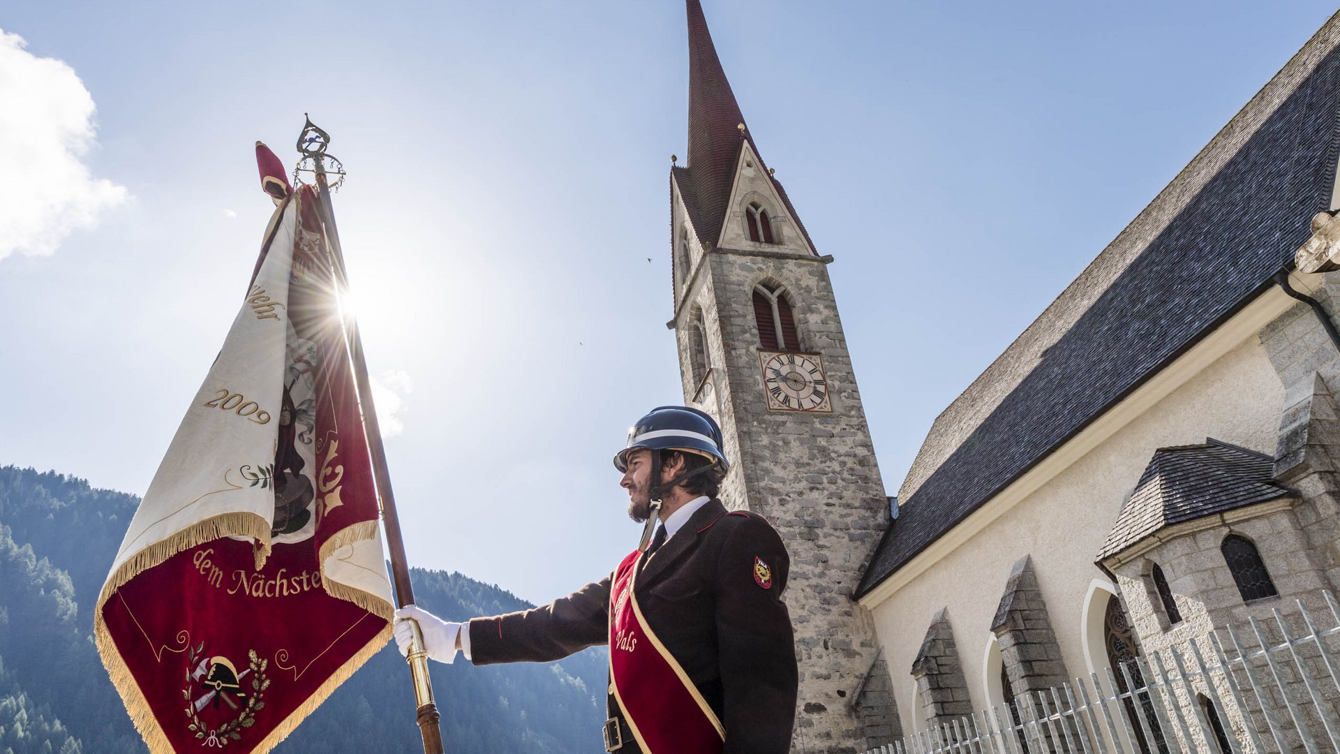 Valles & il villaggio alpino più bello dell’Alto Adige L'immagine mostra un uomo in una divisa tradizionale con elmo che tiene una grande bandiera. Sullo sfondo si vede una chiesa con una torre alta e un orologio, mentre il sole splende dietro la bandiera.