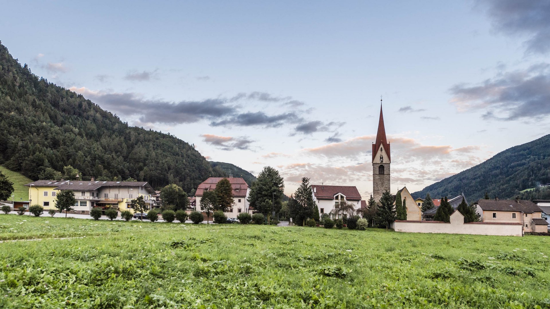 Vandoies: il cuore artigianale della Val Pusteria L'immagine mostra un prato verde in primo piano con una chiesa e diverse case sullo sfondo. La chiesa ha un campanile rosso caratteristico e il villaggio è circondato da colline boschive, mentre il cielo è leggermente nuvoloso.