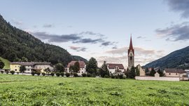 Vandoies: il cuore artigianale della Val Pusteria L'immagine mostra un prato verde in primo piano con una chiesa e diverse case sullo sfondo. La chiesa ha un campanile rosso caratteristico e il villaggio è circondato da colline boschive, mentre il cielo è leggermente nuvoloso.
