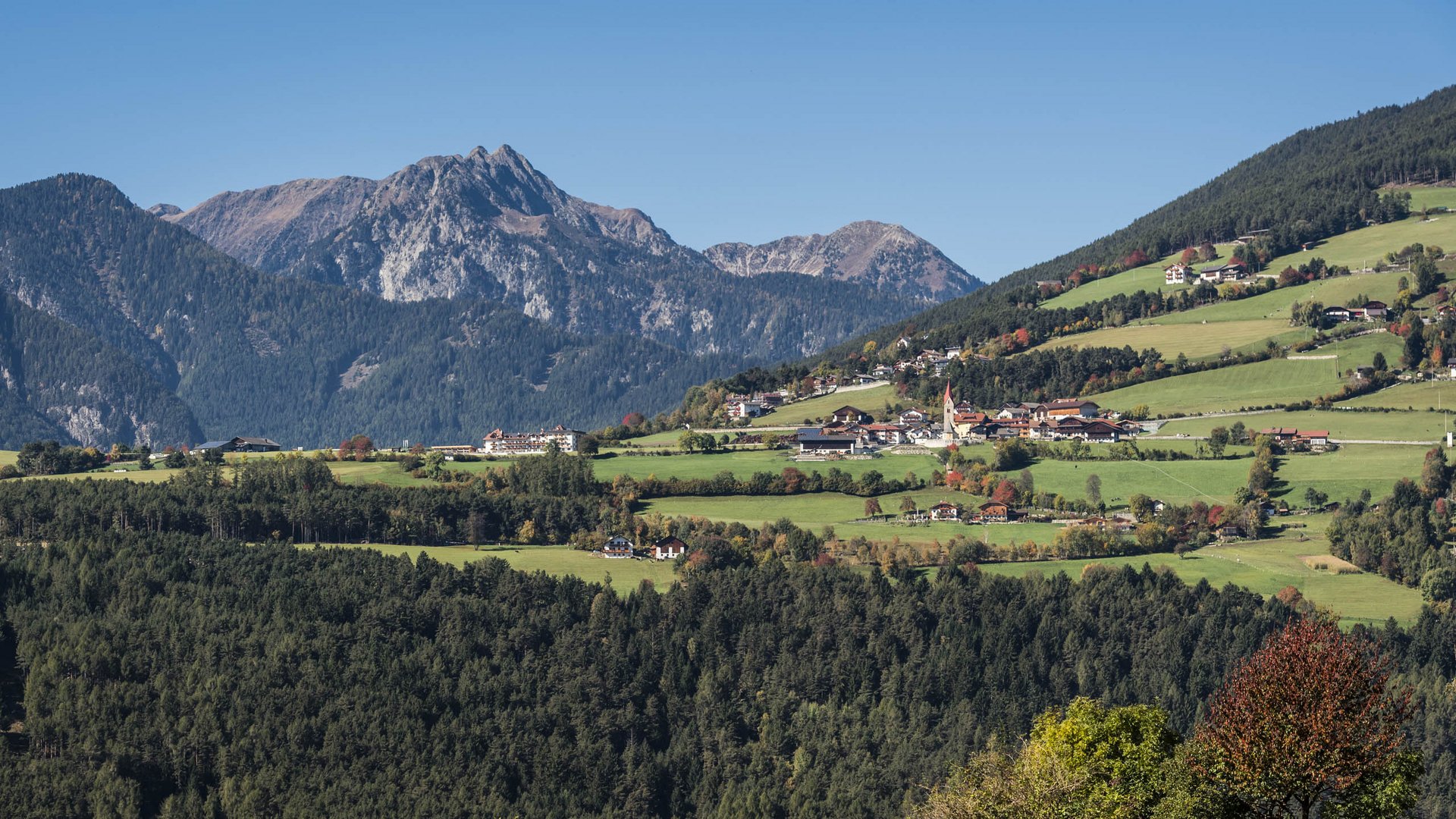La frazione montana di Spinga L'immagine mostra un paesaggio verde e collinoso con case sparse e un campanile al centro. Sullo sfondo si ergono montagne impressionanti che incorniciano la scena.