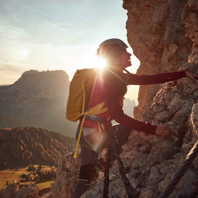 Vacanze estive in Alto Adige: escursioni e altre attività L'immagine mostra una persona che scala una parete rocciosa ripida. Il sole splende direttamente dietro di lei, creando una silhouette suggestiva. La persona indossa un casco e uno zaino giallo, mentre sullo sfondo si vede un vasto paesaggio montano.