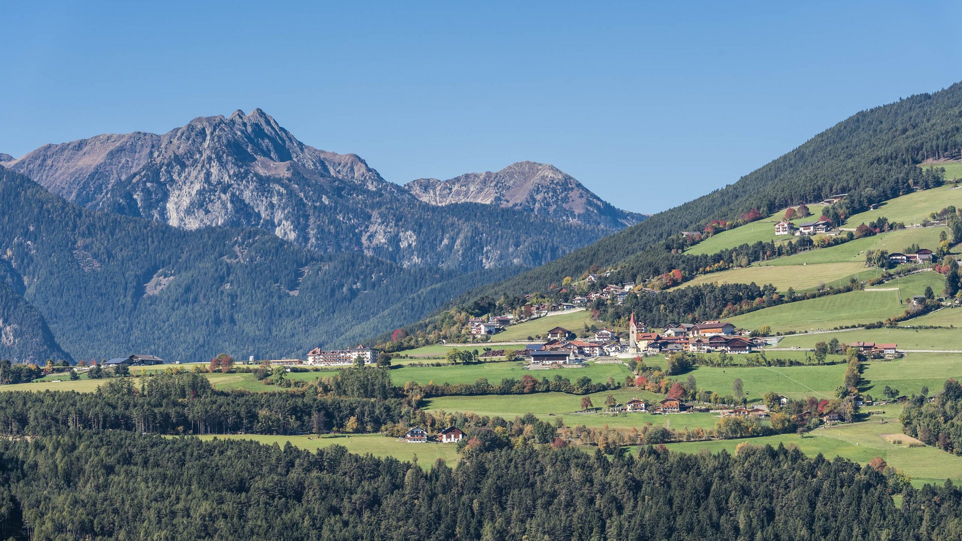 La frazione montana di Spinga L'immagine mostra un paesaggio idilliaco con colline verdi, su cui sono sparse piccole case e una chiesa con una torre rossa. Sullo sfondo si ergono montagne impressionanti, coperte da fitte foreste.