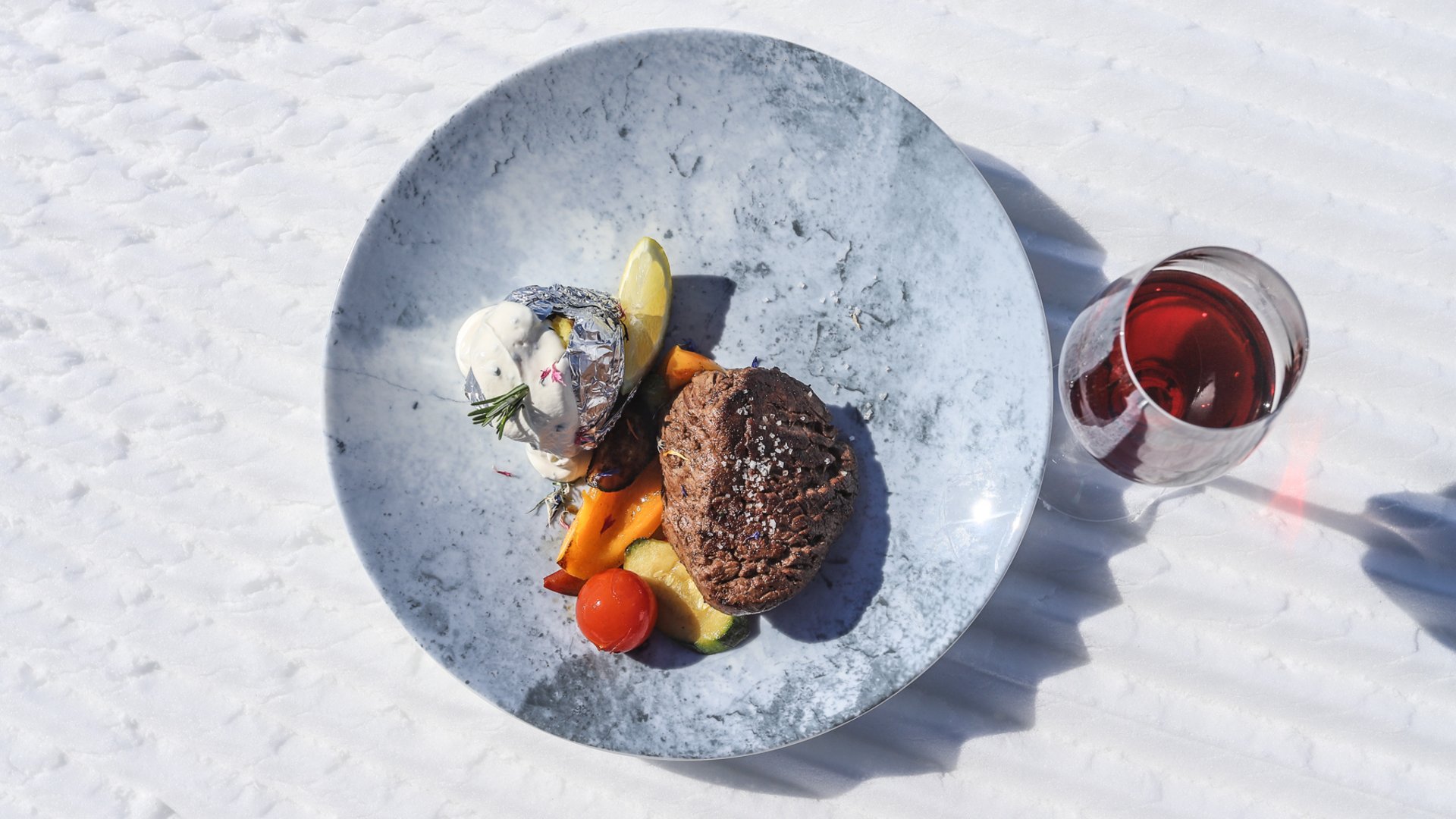Rifugi e malghe di Rio Pusteria Nell'immagine c'è un piatto appetitoso con una bistecca alla griglia, accompagnata da verdure grigliate e una patata al forno avvolta nella carta stagnola con crema alle erbe, servita su un piatto marmorizzato su una superficie coperta di neve accanto a un bicchiere di vino rosso.