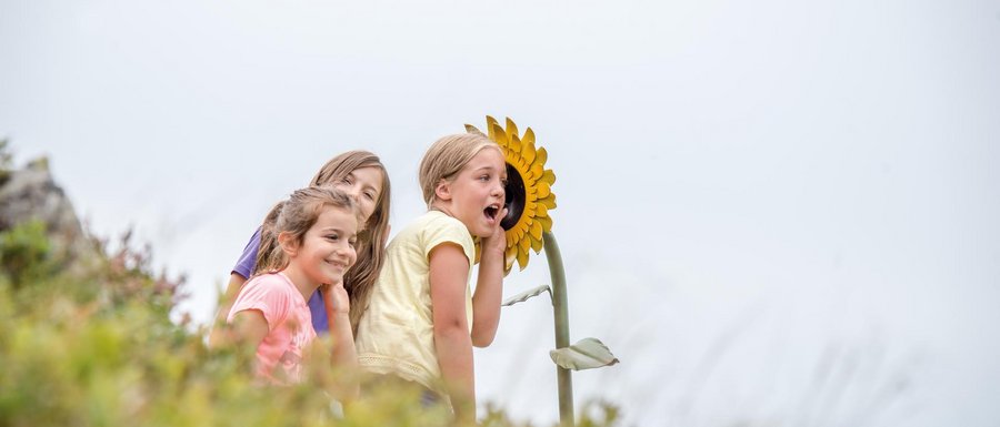 Le località dell’area vacanze Rio Pusteria L'immagine mostra tre bambini sorridenti che si appoggiano a un grande girasole giallo. Il girasole sembra essere parte di un'installazione o di un parco giochi, e i bambini sembrano divertirsi mentre interagiscono con esso.