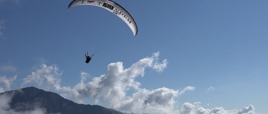 Le località dell’area vacanze Rio Pusteria L'immagine mostra un parapendio che sorvola in alto un paesaggio montano. Il cielo è limpido e azzurro, con alcune nuvole sparse, e si vede la silhouette di una montagna sullo sfondo.