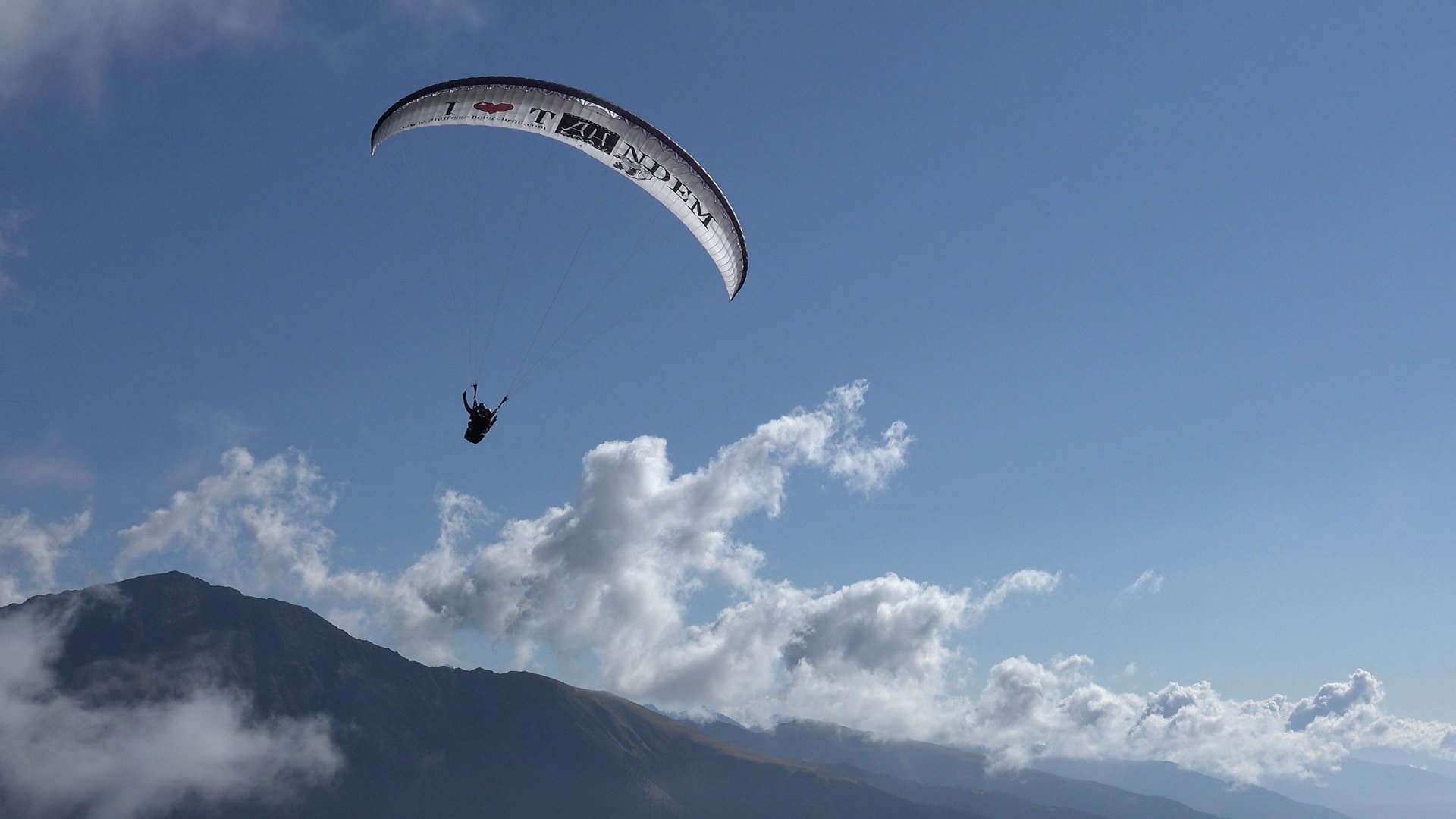 Parapendio in Alto Adige L'immagine mostra un parapendio che sorvola in alto un paesaggio montano. Il cielo è limpido e azzurro, con alcune nuvole sparse, e si vede la silhouette di una montagna sullo sfondo.