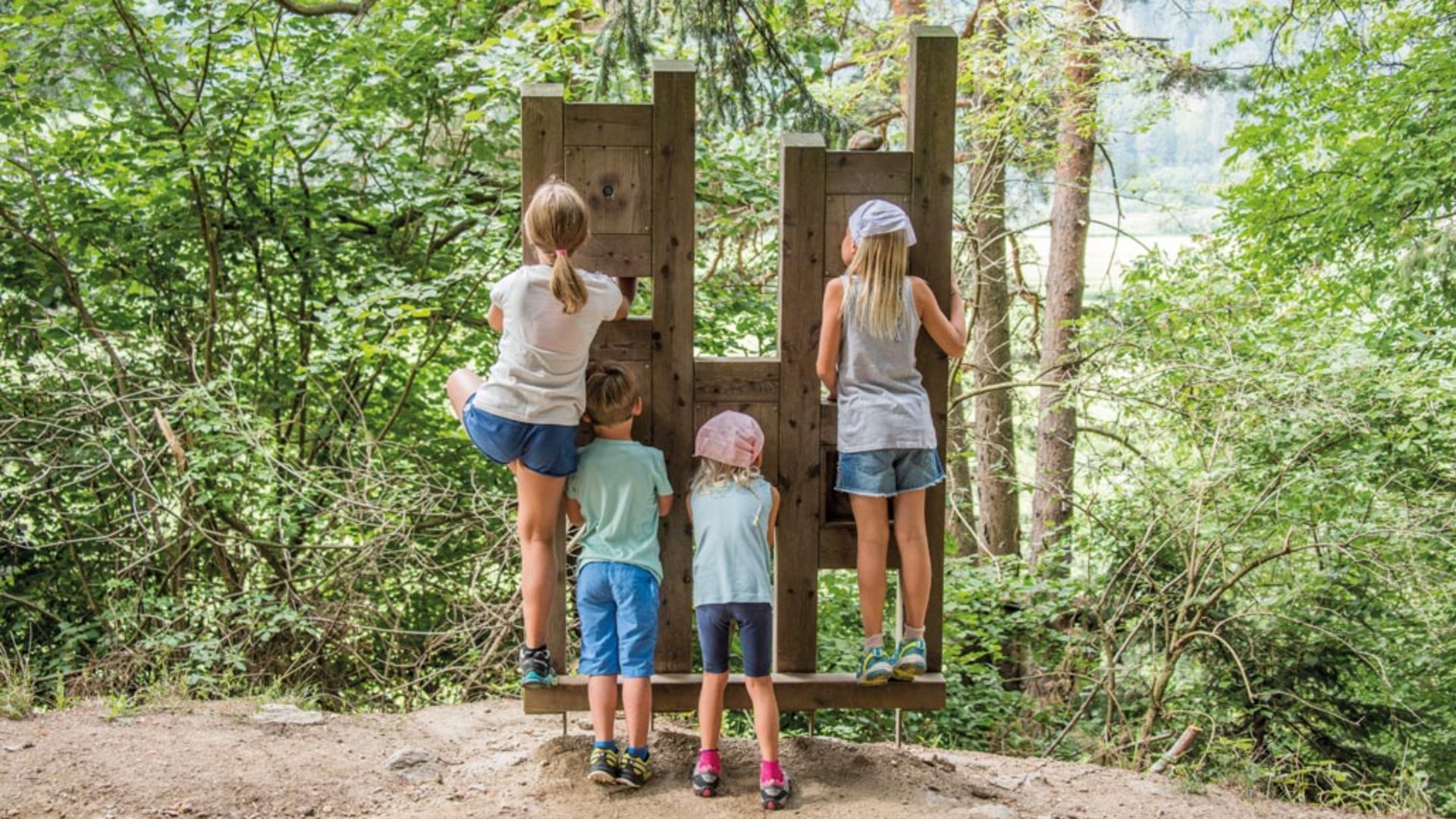 Il Sentiero degli Elfi a Vandoies di Sopra L'immagine mostra quattro bambini che si trovano davanti a una struttura di legno che sembra una sorta di punto panoramico. I bambini guardano attraverso aperture nella struttura verso una foresta verde.