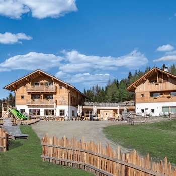 Sapori in malga L'immagine mostra due edifici moderni in legno della "Roner Alm" sotto un cielo azzurro limpido con alcune nuvole bianche sparse. Gli edifici sono circondati da una recinzione in legno e in primo piano si trova un parco giochi con uno scivolo e alcune panchine in legno. Sullo sfondo si vedono foreste e colline.