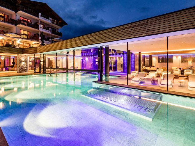 Benvenuti nell’Area Vacanze Sci e Malghe Rio Pusteria! L'immagine mostra una piscina esterna illuminata di notte, circondata da un edificio moderno con grandi pareti di vetro. La piscina si illumina di diversi colori, mentre all'interno si vedono lettini bianchi e un'atmosfera accogliente.