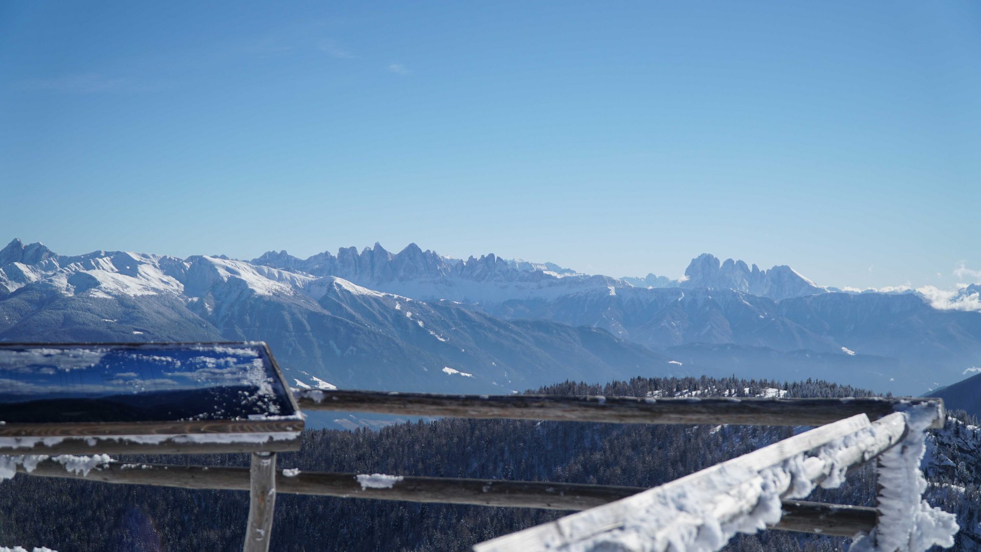 Valles - Jochtal - Steinermandl L'immagine mostra un paesaggio montano invernale con cime innevate sullo sfondo sotto un cielo azzurro intenso. In primo piano si vede una ringhiera coperta di neve e un cartello o una tabella informativa leggermente sfocata.