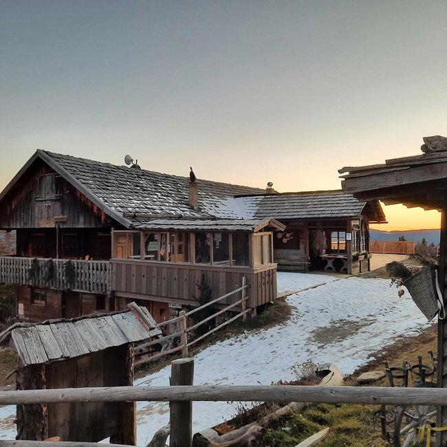 Pertinger Alm L'immagine mostra un rustico chalet di montagna in legno in inverno al tramonto. La baita ha un ampio balcone ed è parzialmente coperta di neve. Sullo sfondo si vedono alberi di conifere e montagne, mentre il sole tramonta all'orizzonte.
