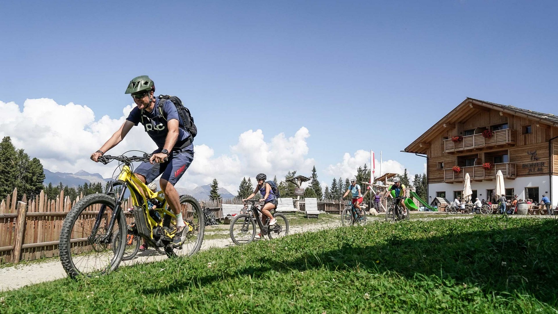 L’Alpe di Rodengo e Luson L'immagine mostra un gruppo di persone che percorrono un sentiero in mountain bike. Sullo sfondo si vede un edificio in legno che sembra un rifugio di montagna, e ci sono altre persone che si trovano lì. Il cielo è limpido e azzurro, e il tempo sembra soleggiato.