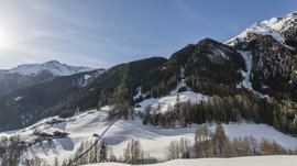 Quando si dice idilliaco… ecco a voi Fundres L'immagine mostra un paesaggio montano coperto di neve con diverse case distribuite sui pendii. Le montagne sullo sfondo sono parzialmente coperte di boschi, e il sole splende in un cielo azzurro e limpido, proiettando lunghe ombre sulla neve.