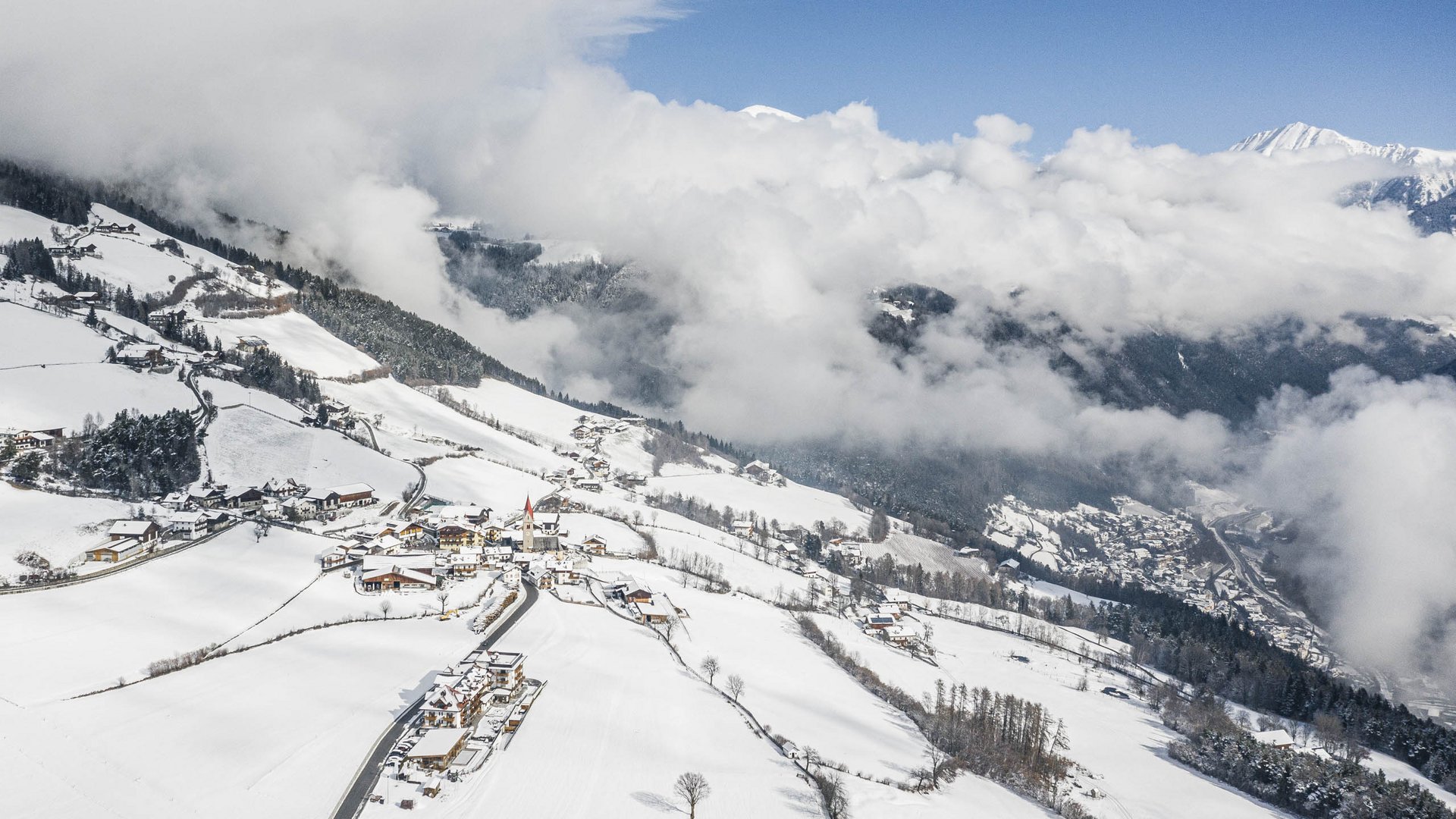 La frazione montana di Spinga L'immagine mostra un villaggio innevato tra le montagne, immerso in paesaggi collinari. Le nuvole avvolgono le valli e le cime circostanti, mentre un cielo azzurro limpido si estende sopra il villaggio.