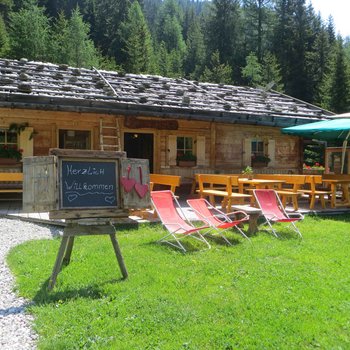 Sapori in malga L'immagine mostra un'affascinante baita di montagna rustica in legno con un cartello di benvenuto "Herzlich Willkommen" davanti, circondata da alberi verdi e un'area relax con tavoli di legno, panche e sdraio sotto un ombrellone.