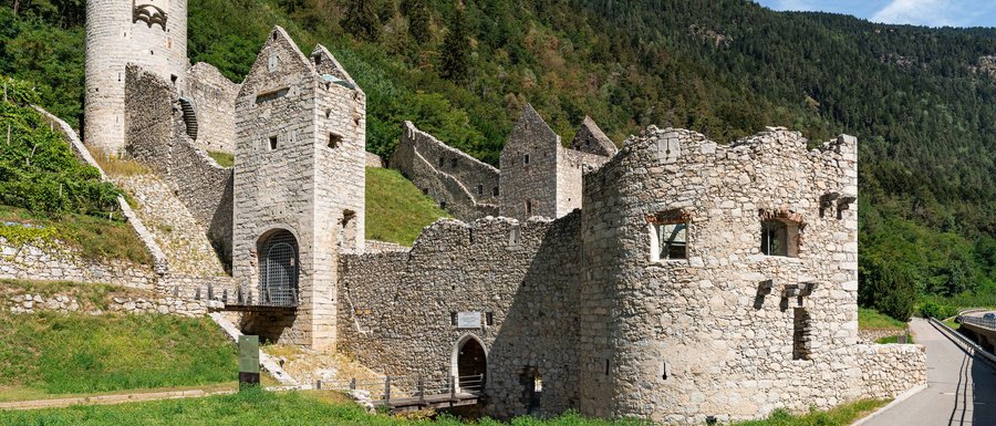 Le località dell’area vacanze Rio Pusteria L'immagine mostra un'antica rovina di un castello in pietra con diverse torri e mura ben conservate. Il castello si trova su un pendio di montagna, circondato da prati verdi e densi boschi, sotto un cielo azzurro limpido.