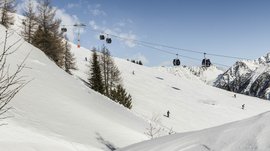 Sciare in Alto Adige: Rio Pusteria L'immagine mostra una pista da sci innevata con diversi sciatori che scendono lungo la pendio. Sopra corre una funivia con cabine che trasporta gli sciatori in cima alla montagna. Sullo sfondo si vedono montagne coperte di neve e un cielo terso e azzurro.