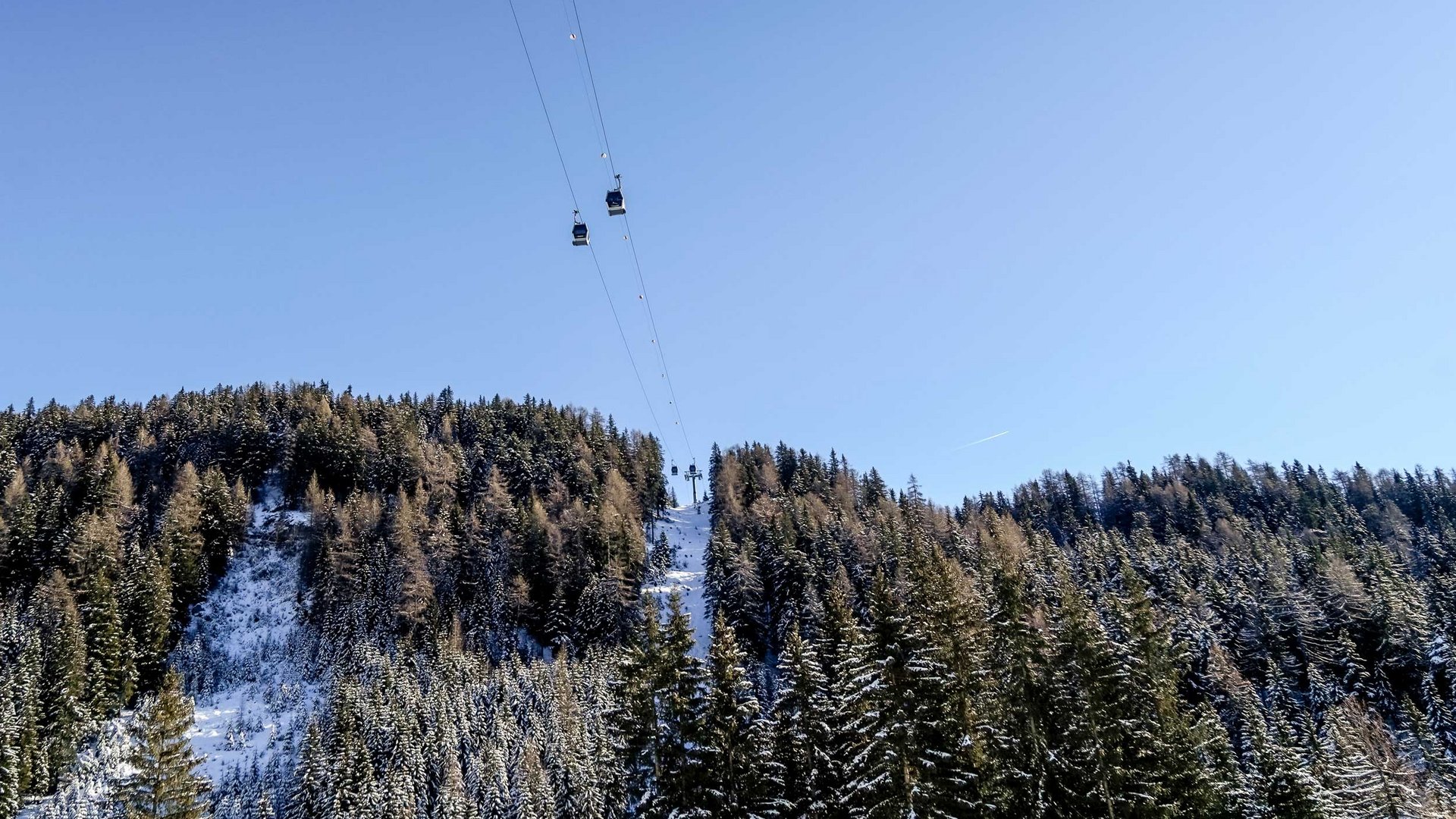 La Valle di Altafossa, presso Maranza L'immagine mostra una funivia con cabine che passano sopra alberi coperti di neve e una collina boscosa. Il cielo è sereno e blu, e la funivia conduce a una cima anch'essa coperta di neve.